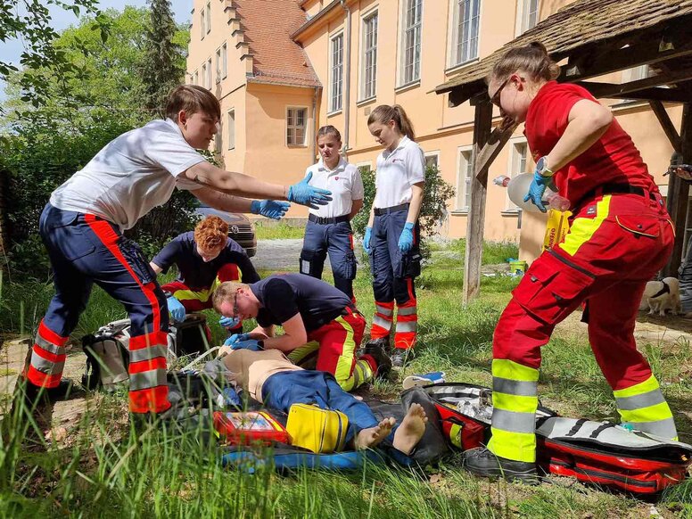 Mehrere Auszubildende im Bereich Notfallmedizin üben die Versorgung einer Person auf dem Boden. Rund um die Übungsszene liegen Rettungsrucksäcke und medizinische Ausrüstung; die praktische Ausbildung findet im Außenbereich eines Gebäudes statt.