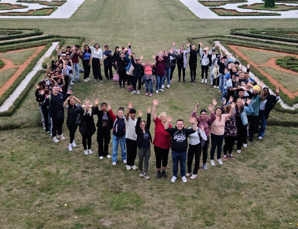 Gruppe von Menschen bildet in einem weitläufigen Schlossgarten eine Herzform und winkt in die Kamera.