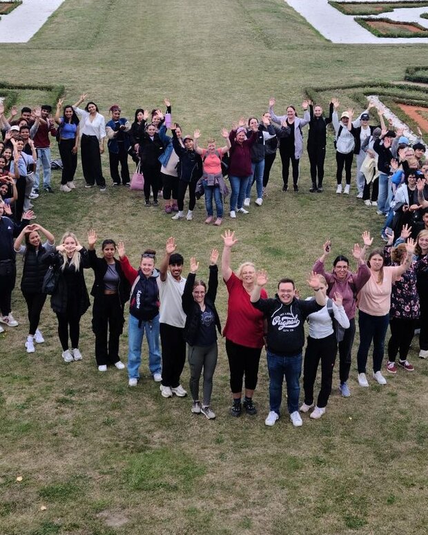 Gruppe von Menschen bildet in einem weitläufigen Schlossgarten eine Herzform und winkt in die Kamera.