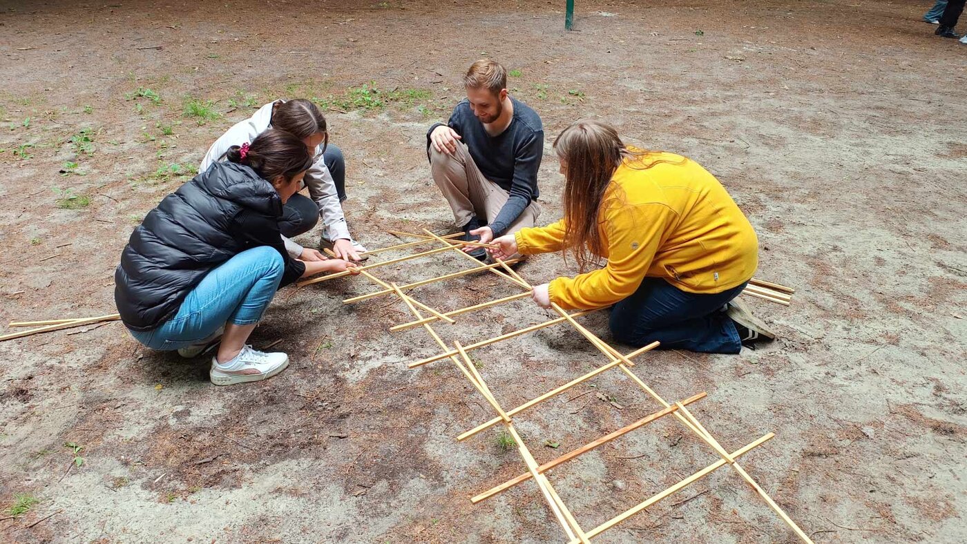 Vier Schüler:innen knien auf einer Waldlichtung und ordnen gemeinsam schmale Holzleisten zu einem gitterartigen Muster auf dem Boden an. Die Gruppe arbeitet an einer kooperativen Aufgabe im Freien.