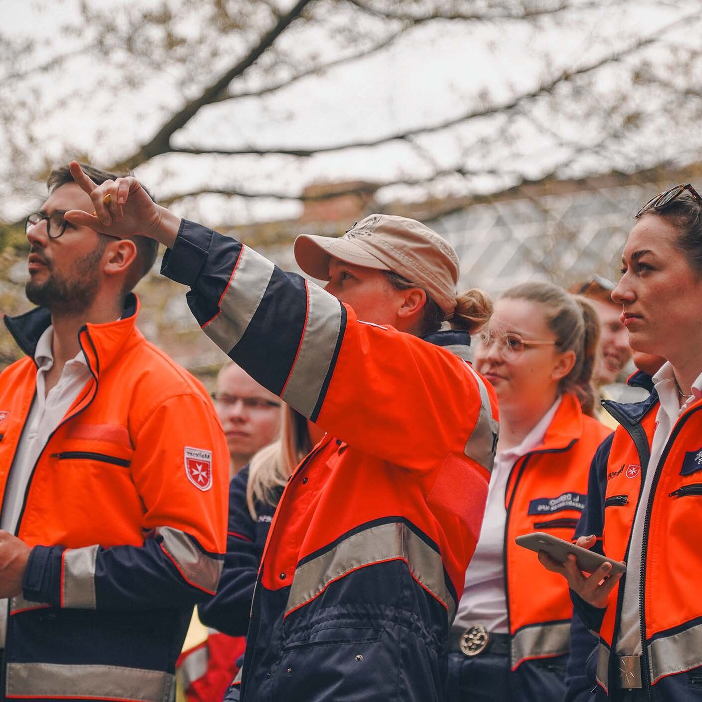 Eine Gruppe von angehenden Rettungskräften in orange-blauer Einsatzkleidung steht im Freien. Eine Person in der Mitte zeigt aufmerksam nach oben, während die anderen in dieselbe Richtung blicken.