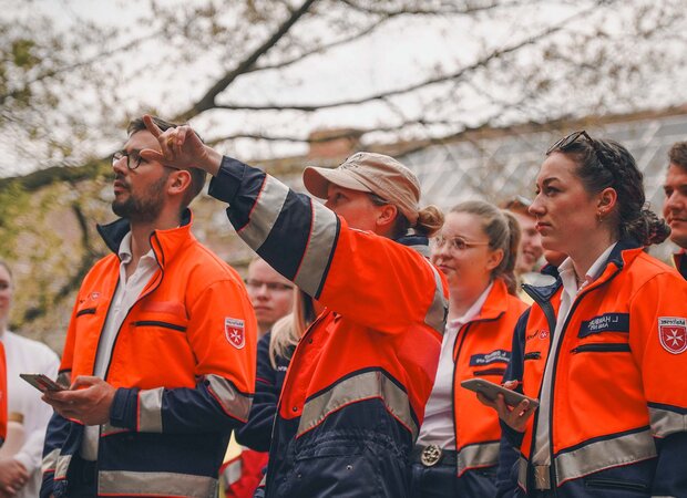 Eine Gruppe von angehenden Rettungskräften in orange-blauer Einsatzkleidung steht im Freien. Eine Person in der Mitte zeigt aufmerksam nach oben, während die anderen in dieselbe Richtung blicken.