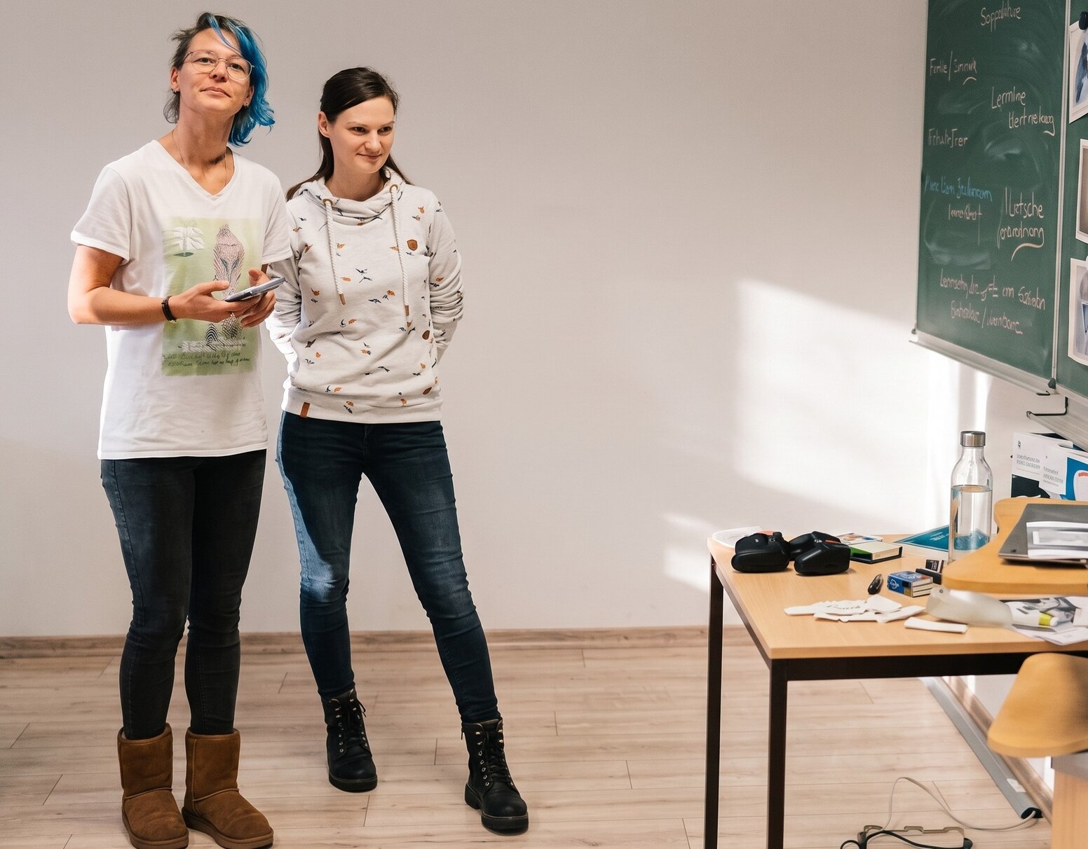 Zwei Personen stehen in einem Unterrichtsraum neben einer Tafel und präsentieren etwas vor einer Gruppe. Auf einem Tisch im Vordergrund liegen Unterrichtsmaterialien, ein Laptop und weitere Arbeitsutensilien.
