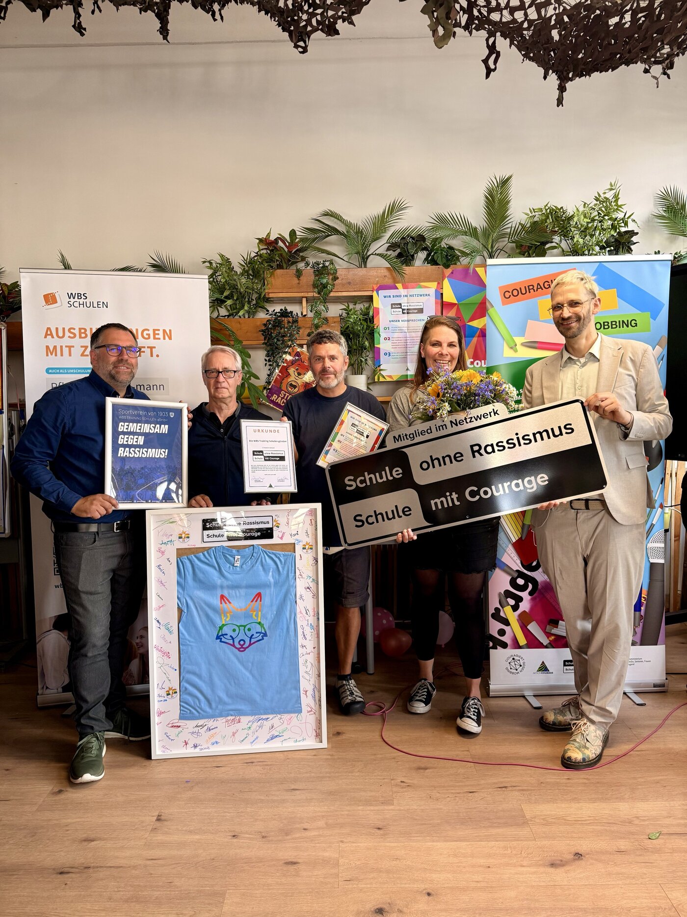 Fünf Mitarbeitende der WBS SCHULEN Berlin präsentieren mehrere Auszeichnungen und Materialien, darunter eine Urkunde, gerahmte Dokumente, ein T-Shirt sowie ein großes Schild mit der Aufschrift „Schule ohne Rassismus – Schule mit Courage“. 