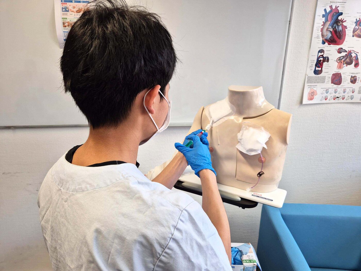 Ein Auszubildender mit Mundschutz und blauen Handschuhen übt an einem medizinischen Torso-Modell die Handhabung einer Spritze.