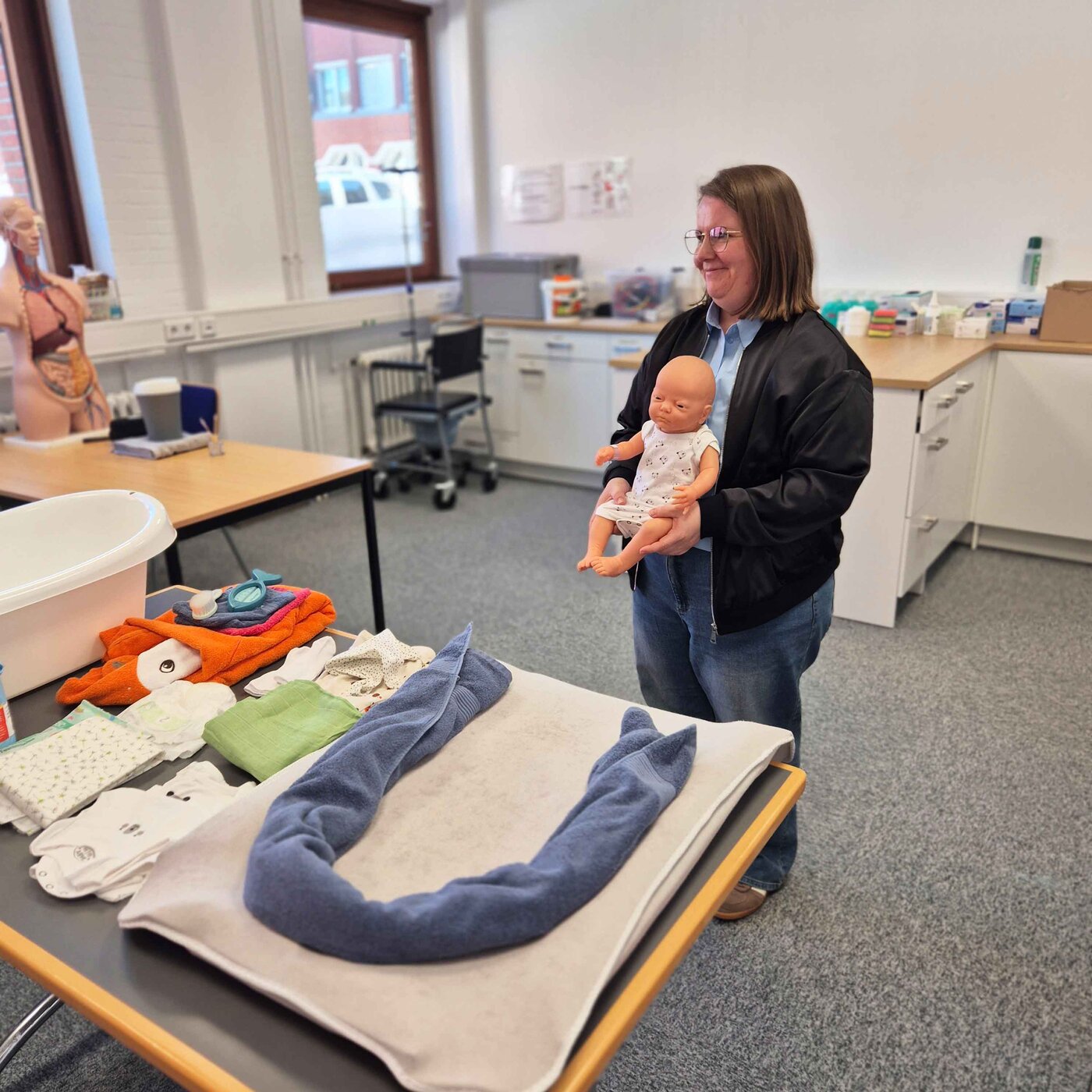 Eine Lernbegleiterin steht Skills Lab und hält eine Babypuppe. Auf dem Tisch davor liegen Pflegeutensilien wie Handtücher, Windeln und eine Babywanne bereit.