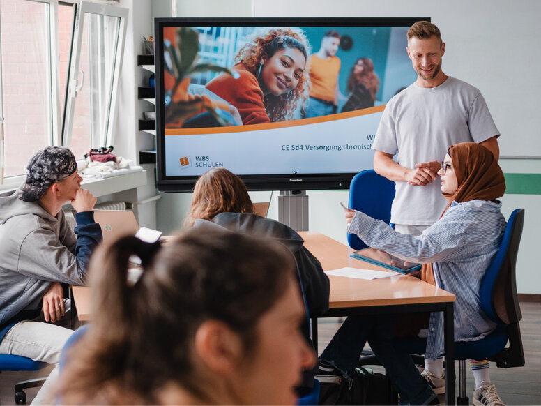 Mehrere Personen sitzen an einem Konferenztisch in einem Schulungsraum und verfolgen eine Präsentation auf einem großen Bildschirm. Auf dem Bildschirm sind das Logo „WBS Schulen“ sowie der Titel „CE 5d4 Versorgung chronisch…“ teilweise sichtbar; eine Lernbegleitung steht neben dem Display und spricht zur Gruppe.