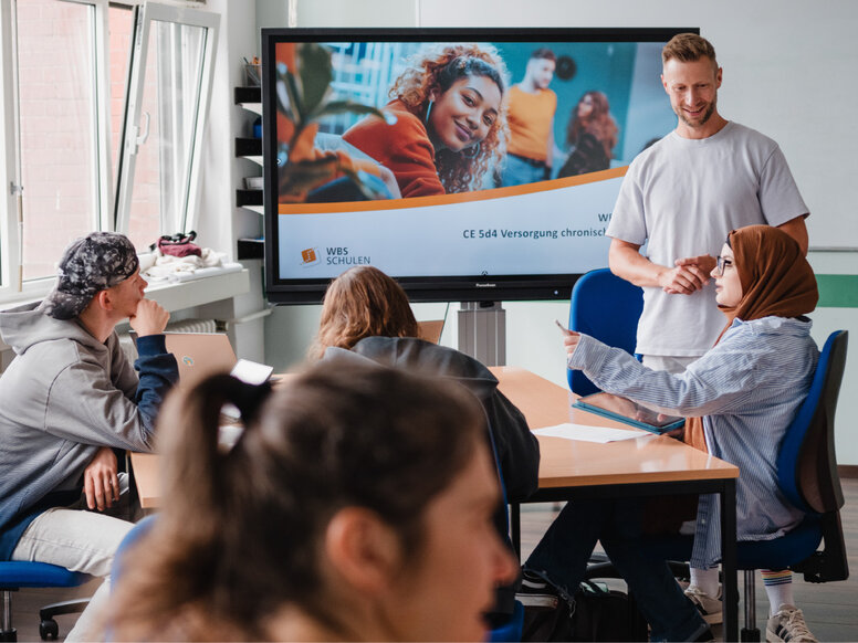 Mehrere Personen sitzen an einem Konferenztisch in einem Schulungsraum und verfolgen eine Präsentation auf einem großen Bildschirm. Auf dem Bildschirm sind das Logo „WBS Schulen“ sowie der Titel „CE 5d4 Versorgung chronisch…“ teilweise sichtbar; eine Lernbegleitung steht neben dem Display und spricht zur Gruppe.