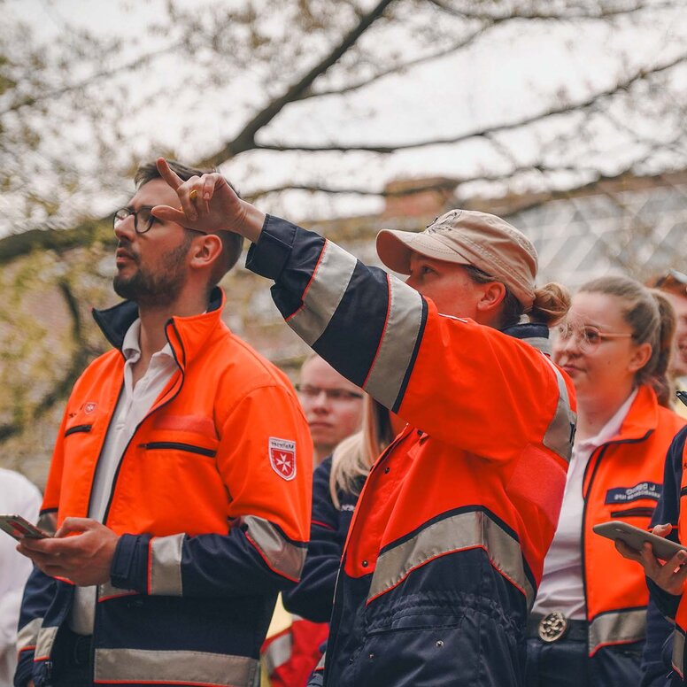 Eine Gruppe von angehenden Rettungskräften in orange-blauer Einsatzkleidung steht im Freien. Eine Person in der Mitte zeigt aufmerksam nach oben, während die anderen in dieselbe Richtung blicken.