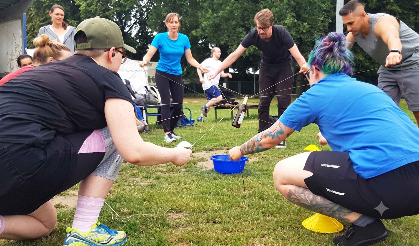 Gruppe bei einer kooperativen Teamchallenge auf einer Wiese. Mit Seilen und Werkzeugen wird gemeinsam eine Aufgabe zur Förderung von Kommunikation gelöst.