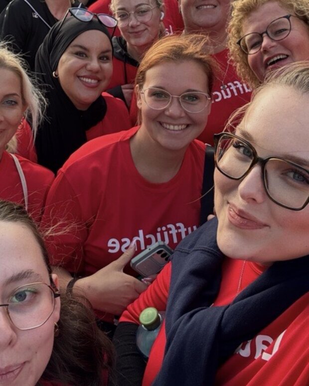 Gruppen-Selfie beim AOK-Firmenlauf in Hamm auf einem Marktplatz: Mehrere Personen in roten T-Shirts stehen dicht vor einer Menschenmenge, im Hintergrund sind weitere Teilnehmende, Fahnen, Stände und historische Gebäude zu sehen.