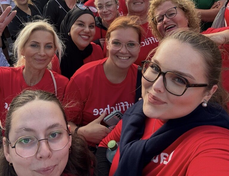 Gruppen-Selfie beim AOK-Firmenlauf in Hamm auf einem Marktplatz: Mehrere Personen in roten T-Shirts stehen dicht vor einer Menschenmenge, im Hintergrund sind weitere Teilnehmende, Fahnen, Stände und historische Gebäude zu sehen.
