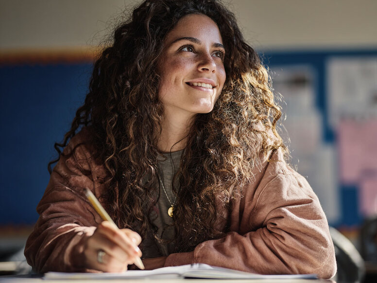 Schülerin sitzt im Klassenzimmer und schaut während einer Lernphase von ihren Notizen hoch. Ihr Blickt schweift in den Raum, während sie lächelt.