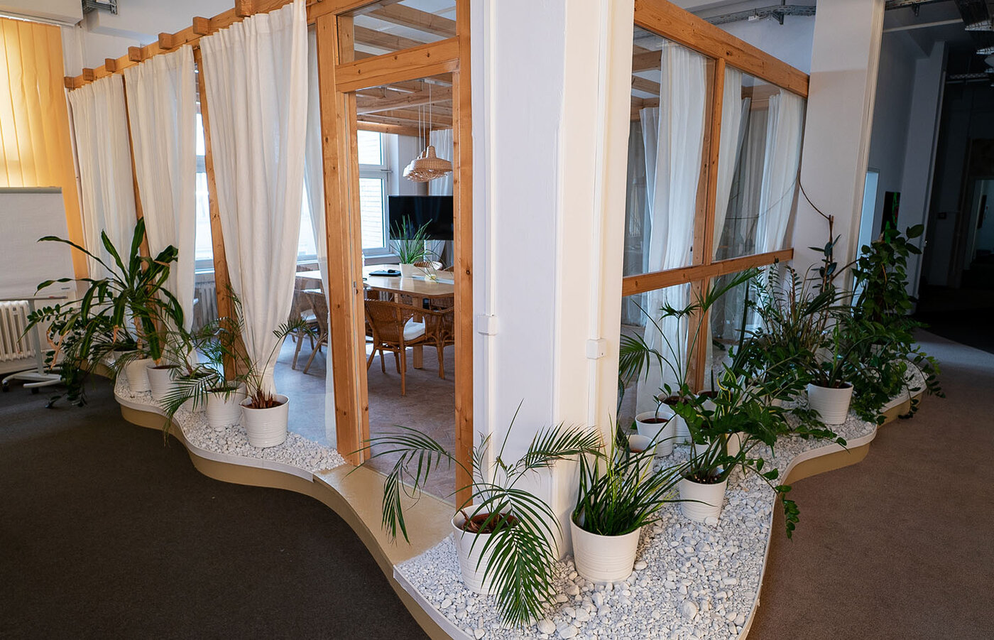 Die Außenansicht eines Besprechungsbereichs innerhalb einer Holzkonstruktion (Pavillon), die in einem größeren Raum steht. Die Konstruktion hat weiße Vorhänge an den Seiten und wird von zahlreichen grünen Topfpflanzen auf weißen Steinen umrandet. Im Inneren ist ein Holztisch mit Korbstühlen zu erkennen.