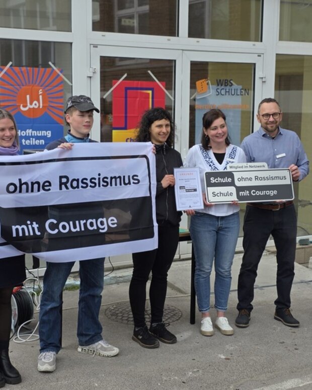 Aufnahmefeier vor einem Schulgebäude: Eine Gruppe präsentiert Banner und Schild „Schule ohne Rassismus – Schule mit Courage“.