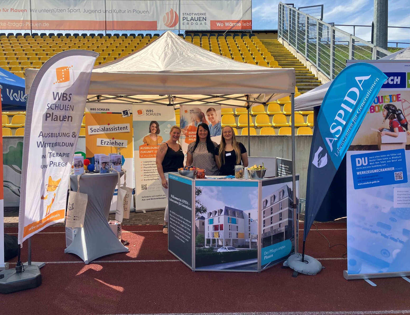 Messestand der WBS SCHULEN Plauen bei einer Ausbildungsmesse im Stadion: Unter einem weißen Pavillon präsentieren drei Personen Informationsmaterial zu Aus- und Weiterbildungen in Therapie, Pflege und Erziehung. Zu sehen sind Roll-ups und Fahnen mit den Hinweisen „Sozialassistent“, „Erzieher“ sowie Werbematerial für Pflegeberufe.