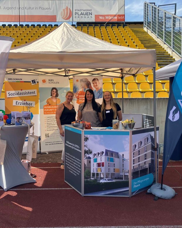 Messestand der WBS SCHULEN Plauen bei einer Ausbildungsmesse im Stadion: Unter einem weißen Pavillon präsentieren drei Personen Informationsmaterial zu Aus- und Weiterbildungen in Therapie, Pflege und Erziehung. Zu sehen sind Roll-ups und Fahnen mit den Hinweisen „Sozialassistent“, „Erzieher“ sowie Werbematerial für Pflegeberufe.