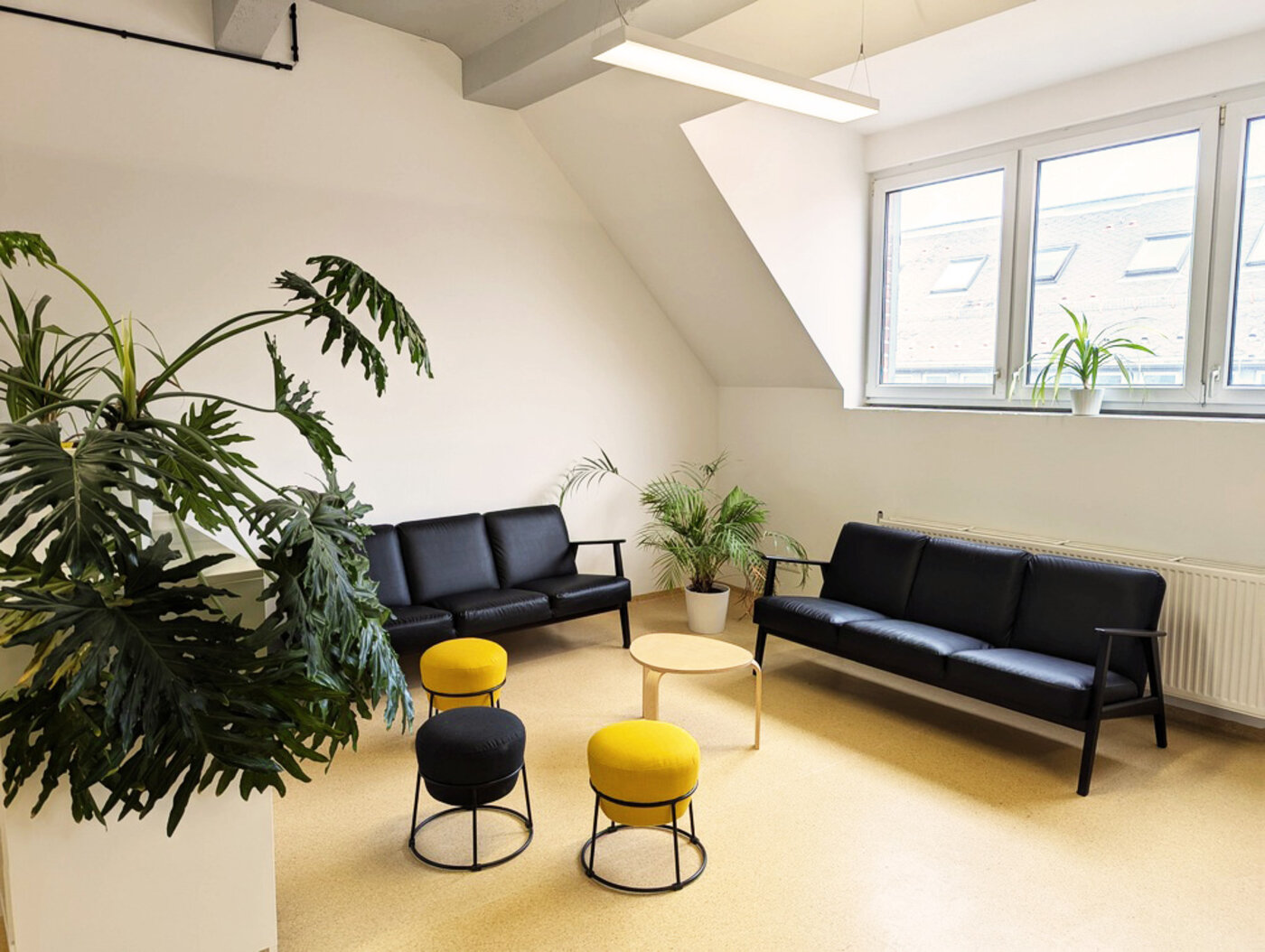 Heller Pausenraum mit schwarzen Sofas, gelben Hockern, Pflanzen und großen Fenstern. Ein ruhiger Bereich zum Entspannen und für Gespräche.