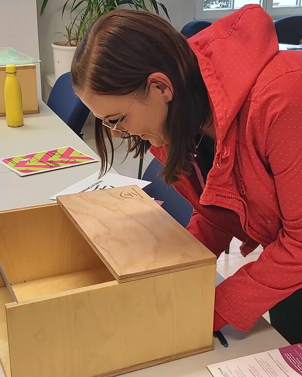 Schülerin in roter Jacke beugt sich über eine geöffnete Holzkiste auf einem Tisch und erkundet deren Inhalt. Die Szene findet in einem hellen Raum mit Tischen, Stühlen und weiteren Holzkisten statt, während weitere Personen im Hintergrund zu sehen sind.