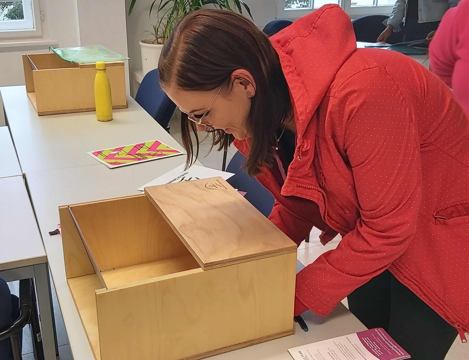 Schülerin in roter Jacke beugt sich über eine geöffnete Holzkiste auf einem Tisch und erkundet deren Inhalt. Die Szene findet in einem hellen Raum mit Tischen, Stühlen und weiteren Holzkisten statt, während weitere Personen im Hintergrund zu sehen sind.