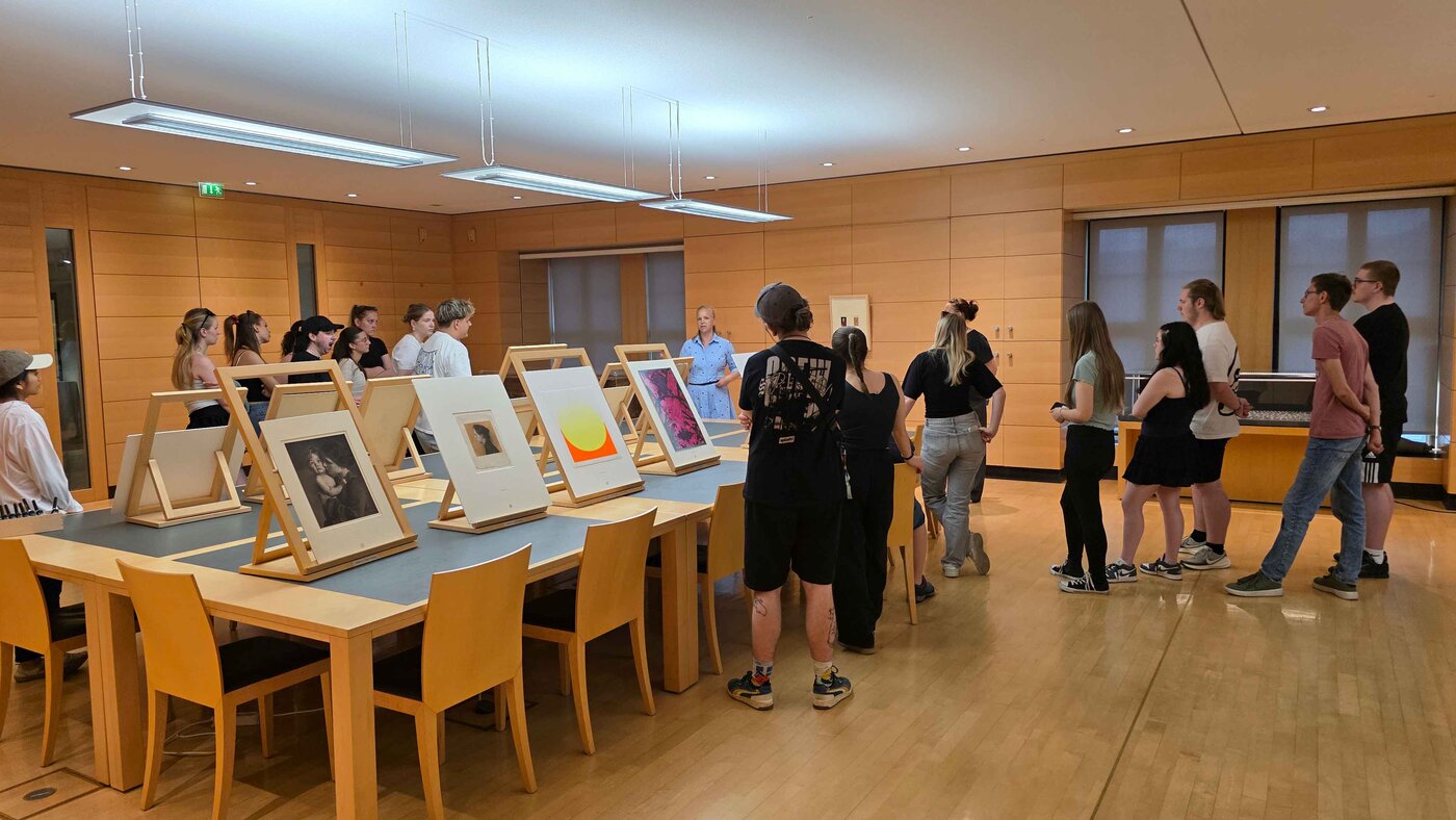 Gruppe von Auszubildenden bei einer Führung in einem Kupferstichkabinett. Die Teilnehmenden stehen um große Tische, auf denen gerahmte Kunstwerke und Drucke auf Holzständern ausgestellt sind.