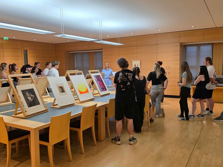 Gruppe von Auszubildenden bei einer Führung in einem Kupferstichkabinett. Die Teilnehmenden stehen um große Tische, auf denen gerahmte Kunstwerke und Drucke auf Holzständern ausgestellt sind.