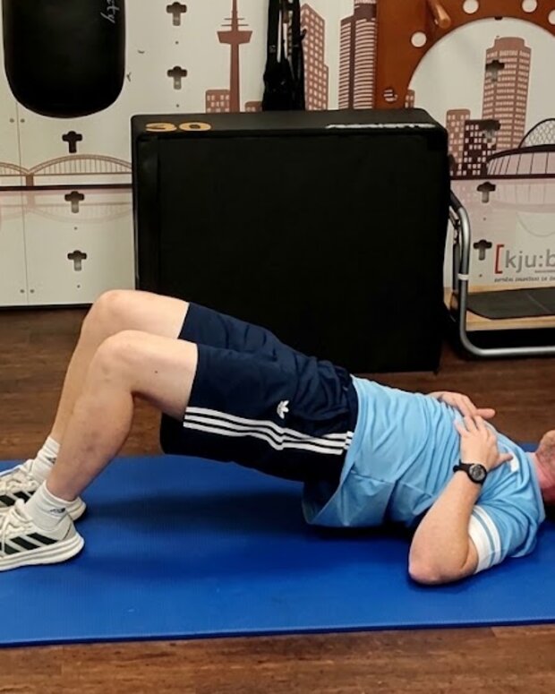 Person führt auf einer Trainingsmatte eine Brückenübung in einem Physiotherapieraum aus.