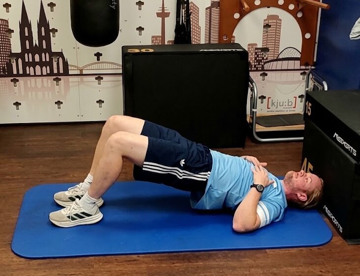 Person führt auf einer Trainingsmatte eine Brückenübung in einem Physiotherapieraum aus.