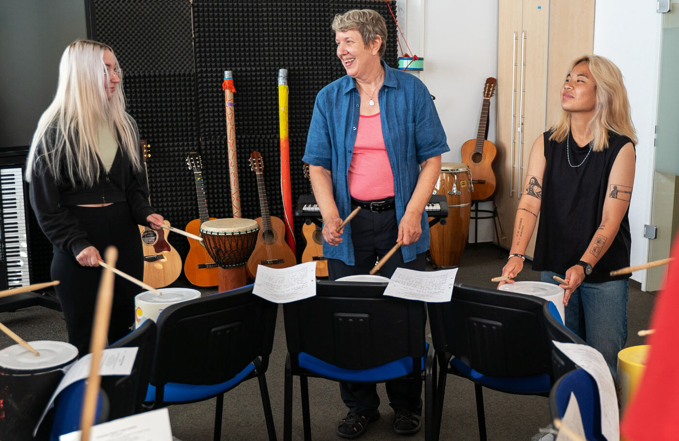 Zwei weibliche Lernende und eine Lernbegleiterin stehen in einem Musikraum. Sie halten Drumsticks in den Händen und stehen vor Stühlen, auf denen umgedrehte weiße Eimer platziert sind. An den Rückenlehnen der Stühle klemmen Notenblätter. Im Hintergrund stehen verschiedene Instrumente wie Gitarren, ein Keyboard und Trommeln vor mit Akustikschaumstoff verkleideten Wänden.