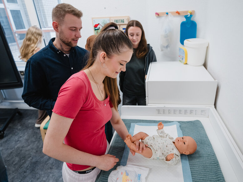 Mehrere Personen beobachten eine praktische Übung zur Babypflege. Im Vordergrund wird eine Babypuppe auf einer Wickelstation versorgt.