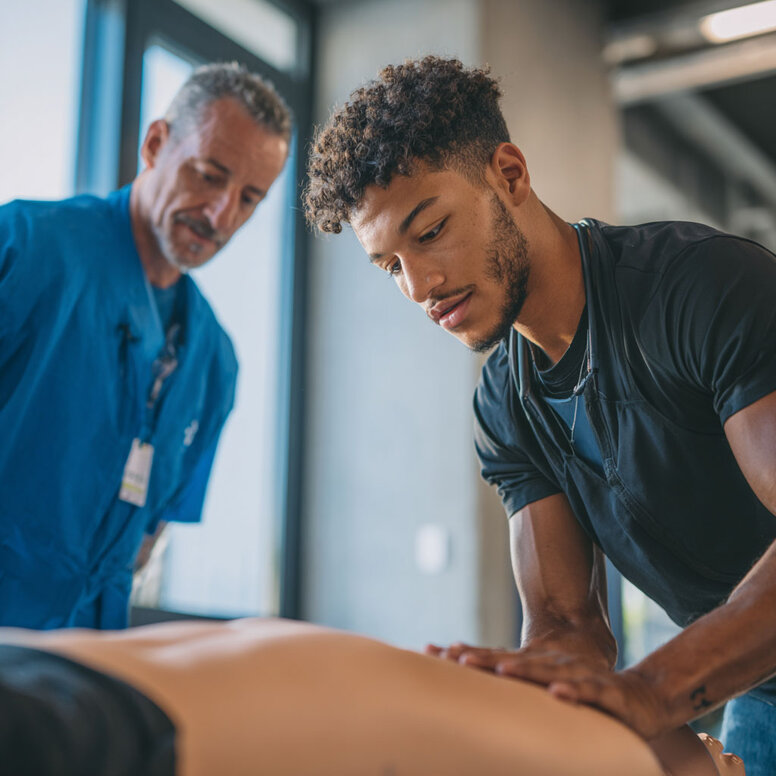 Auszubildender zum Physiotherapeuten übt Massagetechniken an einem Patienten in einer Praxis. Ein erfahrerener Praxisanleiter schaut ihm über die Schulter und gibt Tipps.