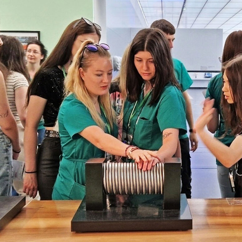 Gruppe junger Frauen, teils in grünen Kasacks, steht in einem Museum um ein wissenschaftliches Exponat aus Metall und berührt dieses.