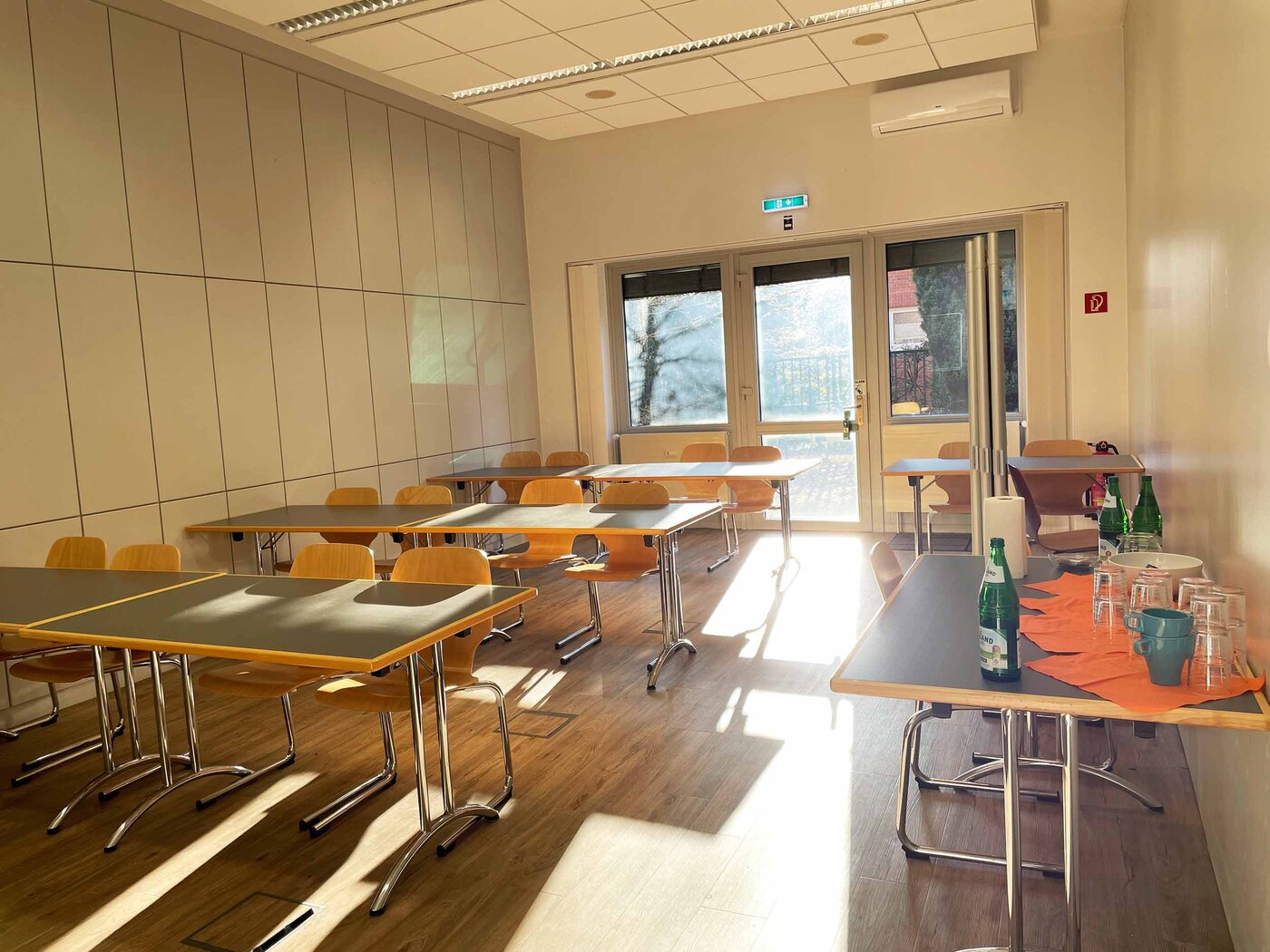 Ein heller, leerer Unterrichtsraum mit Tischen und Holzstühlen am WBS SCHULEN Standort Leer. Durch eine Glastür im Hintergrund fällt Sonnenlicht auf den Boden.