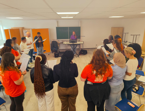 Eine Gruppe von etwa 20 Jugendlichen und jungen Erwachsenen steht in einem Stuhlkreis in einem Klassenraum und hält Notenblätter in den Händen. In der Mitte begleitet eine Person am E-Piano den gemeinsamen Gesang; im Hintergrund sind eine Tafel, Stühle und Unterrichtsmobiliar zu sehen.