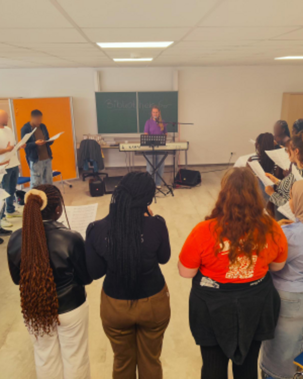 Eine Gruppe von etwa 20 Jugendlichen und jungen Erwachsenen steht in einem Stuhlkreis in einem Klassenraum und hält Notenblätter in den Händen. In der Mitte begleitet eine Person am E-Piano den gemeinsamen Gesang; im Hintergrund sind eine Tafel, Stühle und Unterrichtsmobiliar zu sehen.