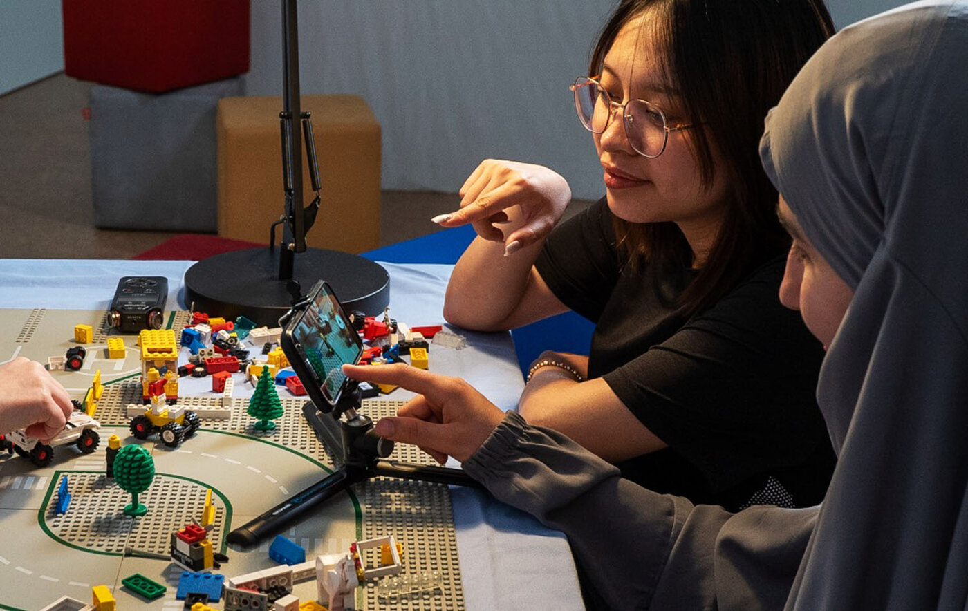 Zwei weibliche Lernende sitzen an einem Tisch und schauen konzentriert auf ein Smartphone, das auf einem kleinen Stativ befestigt ist. Eine der Lernenden zeigt mit dem Finger auf das Display. Auf dem Tisch ist eine Szenerie aus Legosteinen, Spielfiguren und kleinen Spielzeugautos aufgebaut. Daneben liegt ein Audio-Aufnahmegerät.