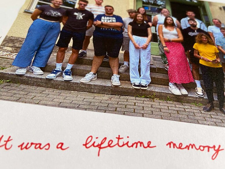 Seite aus einem Erasmus+-Gästebuch mit dem Gruppenfoto von jungen Menschen auf einer Treppe und dem handschriftlichen roten Text "It was a lifetime memory!".