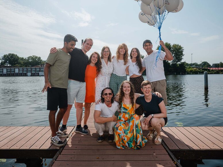 Eine Gruppe Mitarbeitender der WBS GRUPPE steht auf einem Steg an der Havel in Berlin und blicken lachend in die Kamera. Einige umarmen sich, eine Person hält Luftballons in der Hand.