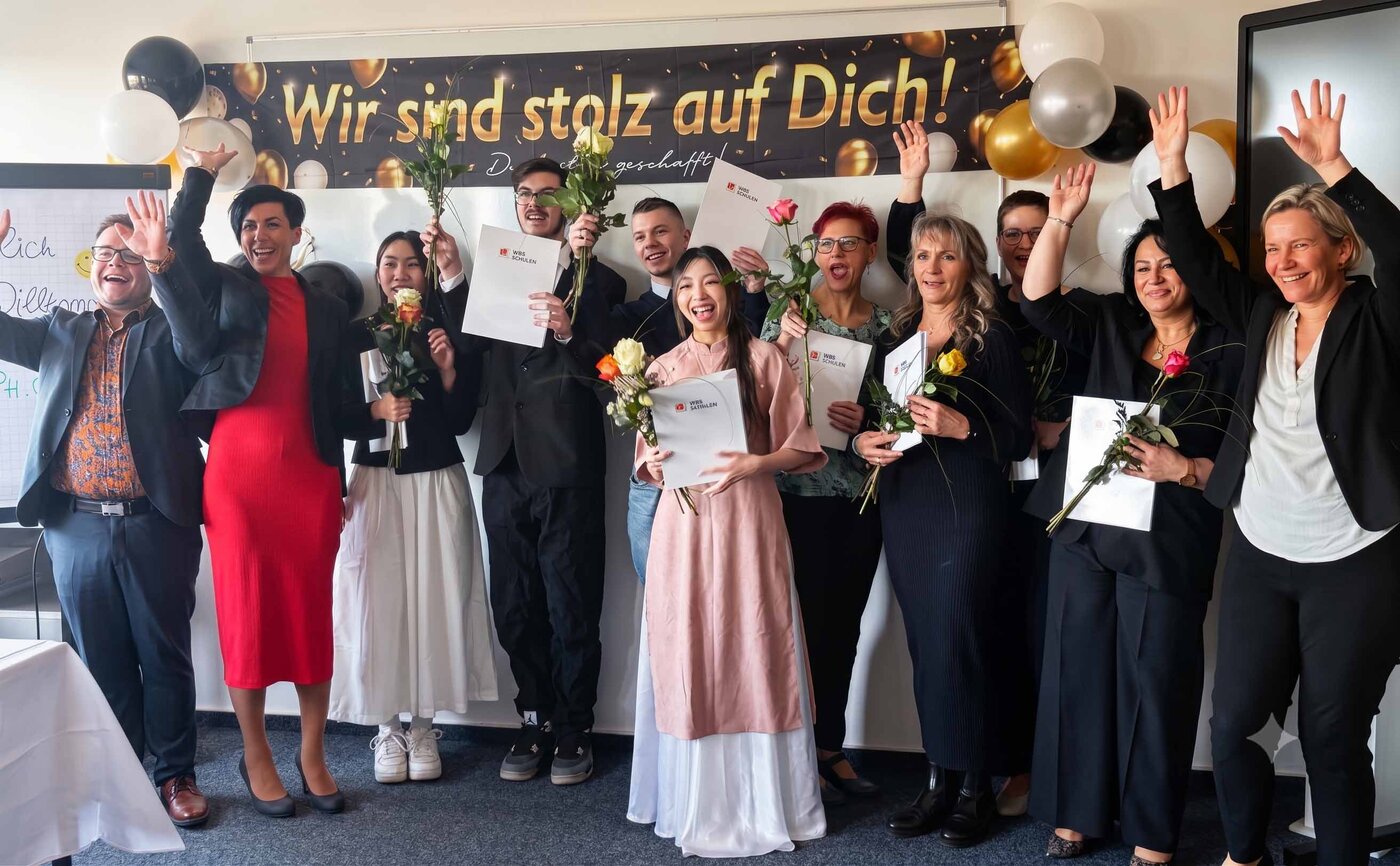 Gruppenfoto einer feierlichen Zeugnisübergabe. Neun glückliche Absolvent:innen und Lernbegleiter:innen stehen vor einem Banner mit der Aufschrift „Wir sind stolz auf Dich! Du hast es geschafft!“, umgeben von Luftballons. Die Personen halten Blumen und Urkunden in den Händen und winken freudig in die Kamera.