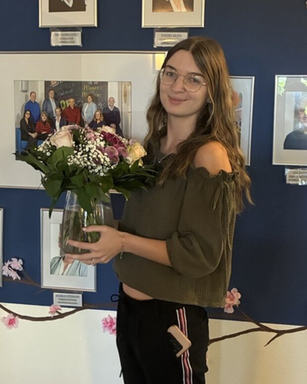 Neue Lernbegleitung mit Blumenstrauß vor einer Fotowand der Mitarbeitenden der WBS SCHULEN.
