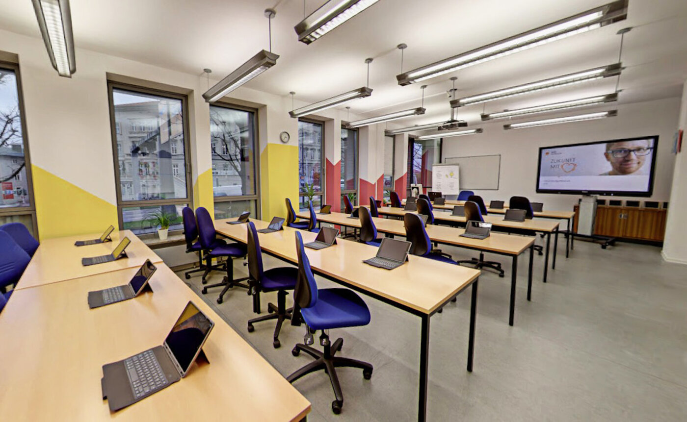 Ein modernes, leeres Klassenzimmer mit großen Fenstern. Auf den holzfarbenen Tischen stehen Tablets bereit, vorn hängt ein großes Smartboard.