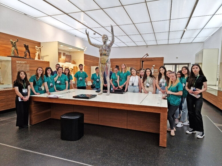 Große Gruppe von Lernenden in grünen Kasacks steht in einem Museum um einen großen Tisch mit einem anatomischen Modell eines Menschen.