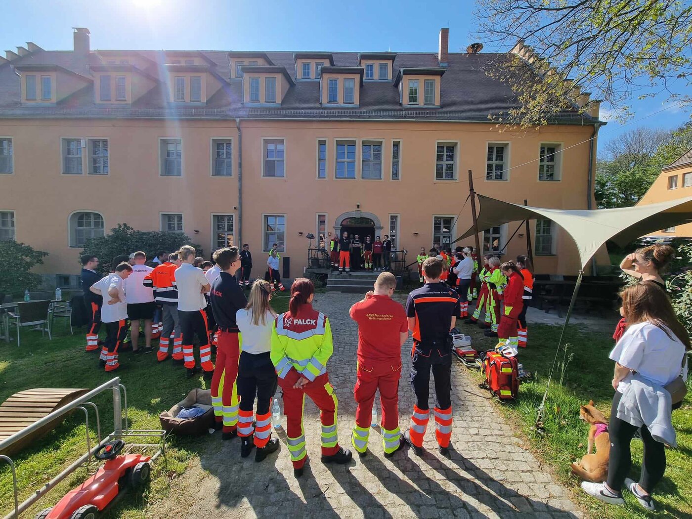 Gruppe von Personen in Rettungsdienstkleidung versammelt sich vor einem größeren Gebäude zu einer Lagebesprechung im Freien. Auf dem Weg vor dem Gebäude stehen Ausrüstungsgegenstände für eine praktische Übung bereit.