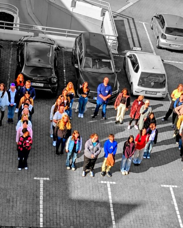 Luftbild eines Parkplatzs mit einer größeren Gruppe von Personen zwischen geparkten Fahrzeugen; die Menschen sind farbig dargestellt, der Rest des Bildes in Schwarz-Weiß. Sie formen das Wort MOIN.