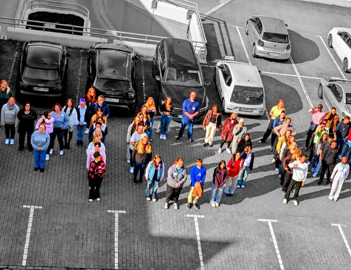 Luftbild eines Parkplatzs mit einer größeren Gruppe von Personen zwischen geparkten Fahrzeugen; die Menschen sind farbig dargestellt, der Rest des Bildes in Schwarz-Weiß. Sie formen das Wort MOIN.