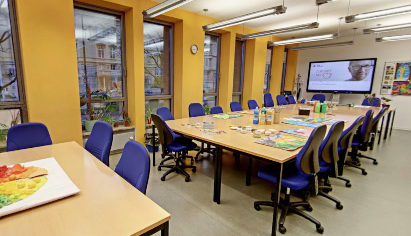 Ein helles Klassenzimmer mit gelben Wänden. Auf einem großen Tisch mit blauen Stühlen liegen bunte Farben, Pinsel und gemalte Bilder bereit.