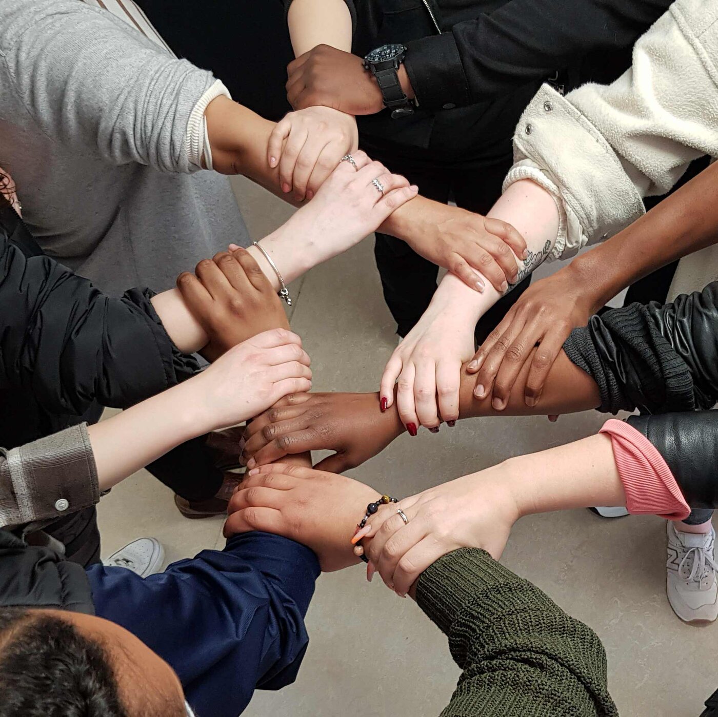 Mehrere Personen stehen im Kreis und halten die Handgelenke der jeweils benachbarten Person fest. Die Aufnahme zeigt eine gemeinsame Team- oder Vertrauensübung, bei der ein geschlossener Verbund aus Armen entsteht.
