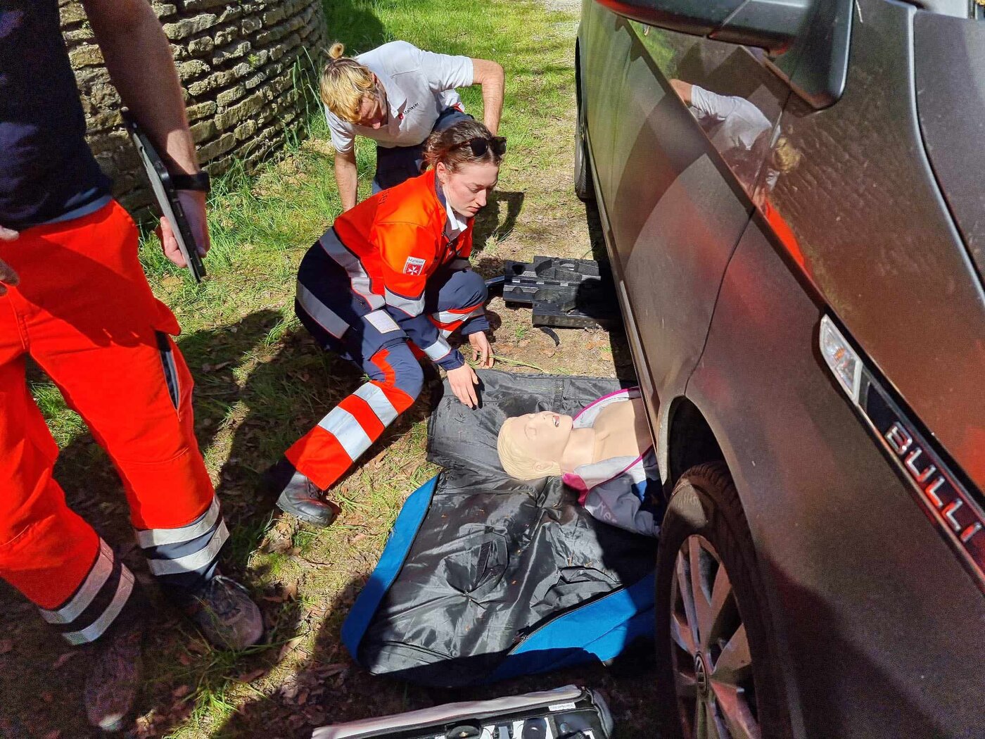 Mehrere Personen in Rettungsdienstkleidung üben an einem Fahrzeug die Versorgung einer eingeklemmten Person. Eine Übungspuppe liegt teilweise unter dem Auto, während die Gruppe die Rettungssituation absichert und beurteilt.