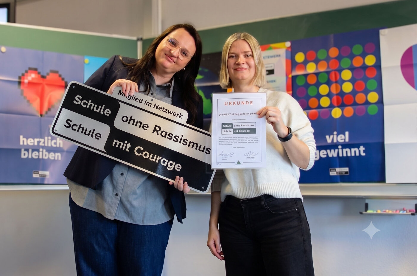 Zwei lächelnde Frauen stehen vor einer Tafel. Die linke hält ein großes Schild mit der Aufschrift „Schule ohne Rassismus – Schule mit Courage“, die rechte präsentiert stolz die dazugehörige Urkunde.