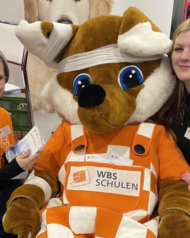 Zwei Auszubildende mit einem Fuchs-Maskottchen am Messestand der WBS SCHULEN bei einer Berufsorientierungsmesse.